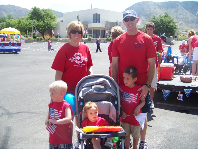 military family wearing red shirts. man and woman. 2 boys and 1 girl sitting in a stroller in front of a Independence Day parade float.