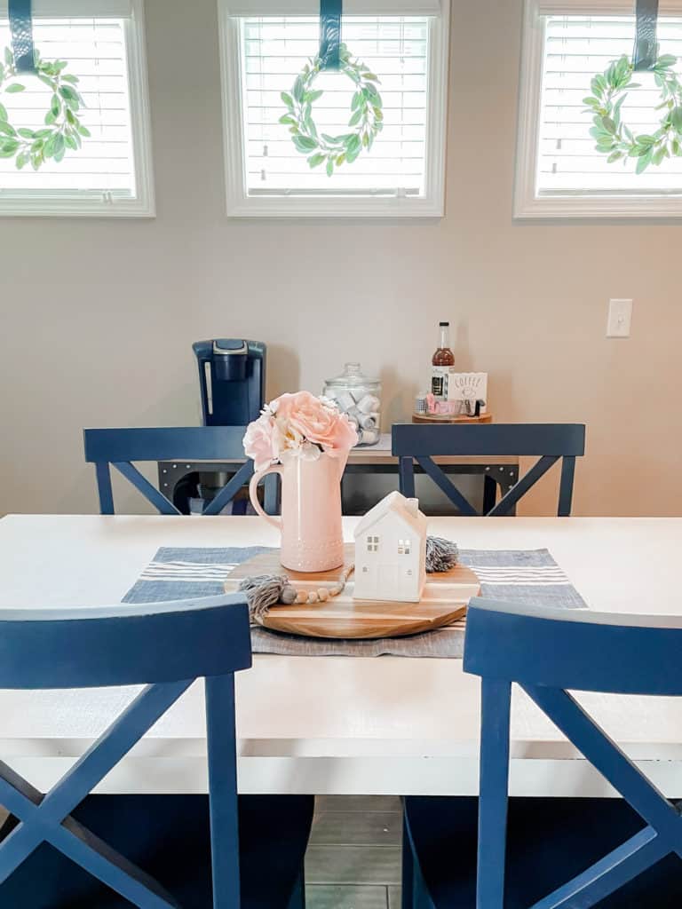 Tidy home tips and a tidy white table with blue chairs. 3 square windows in the background. centerpiece of round wood piece with pink vase and pink flowers and a small white ceramic house.