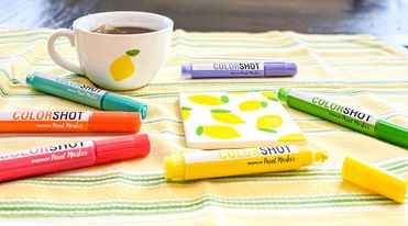 yellow and green placemat with green, orange, pink, purple, and yellow paint markers scattered. white tea cup with a lemon painted on the front of it and a coaster with lemons in the center.