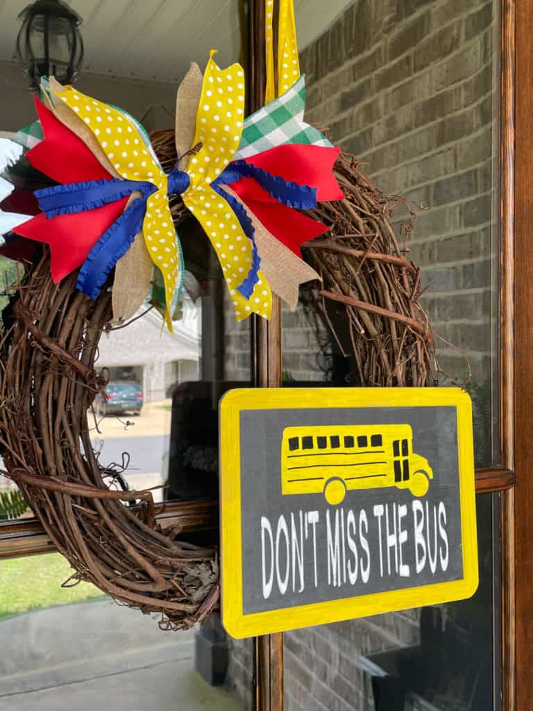 Grapevine wreath with colorful bow and rectangular chalkboard sign that has a yellow school bus and says don't miss the bus.