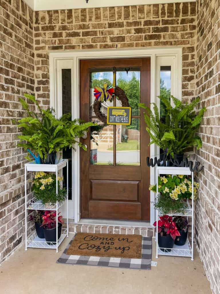 Brown, wood front door with window in the center. wreath with colorful bow hanging in center. flowers and plants in white plant stands on right and left of door.