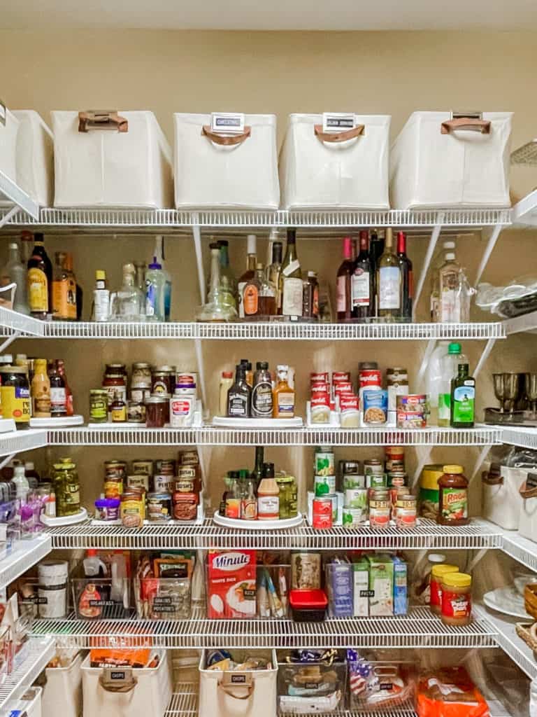 large kitchen pantry with wire shelves. off white canvas bins on top shelves, bottles of alcohol on turntables, canned goods, boxes and other food items organized on shelf risers, turntables and clear plastic containers