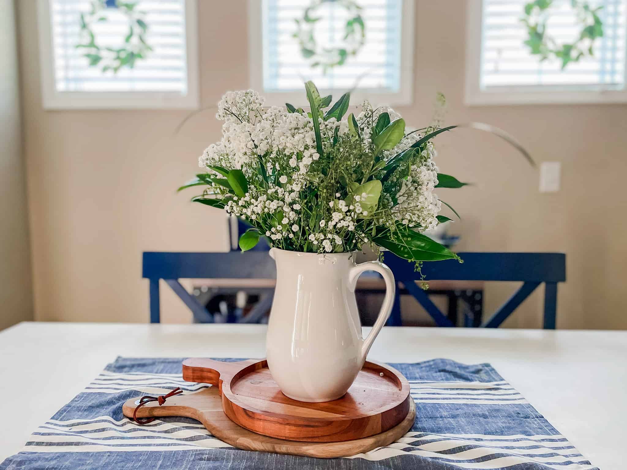 bouquet of greenery and baby's breath in white pitcher sitting on 2 wood cutting boards on a blue and white striped cloth. blue chairs and small windows with wreaths in the background