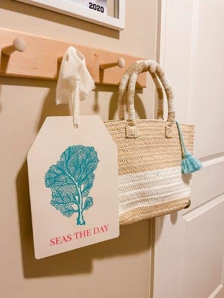 Light wood sign with blue painting and pink writing that says seas the day hanging beside natural jute purse with blue tassel