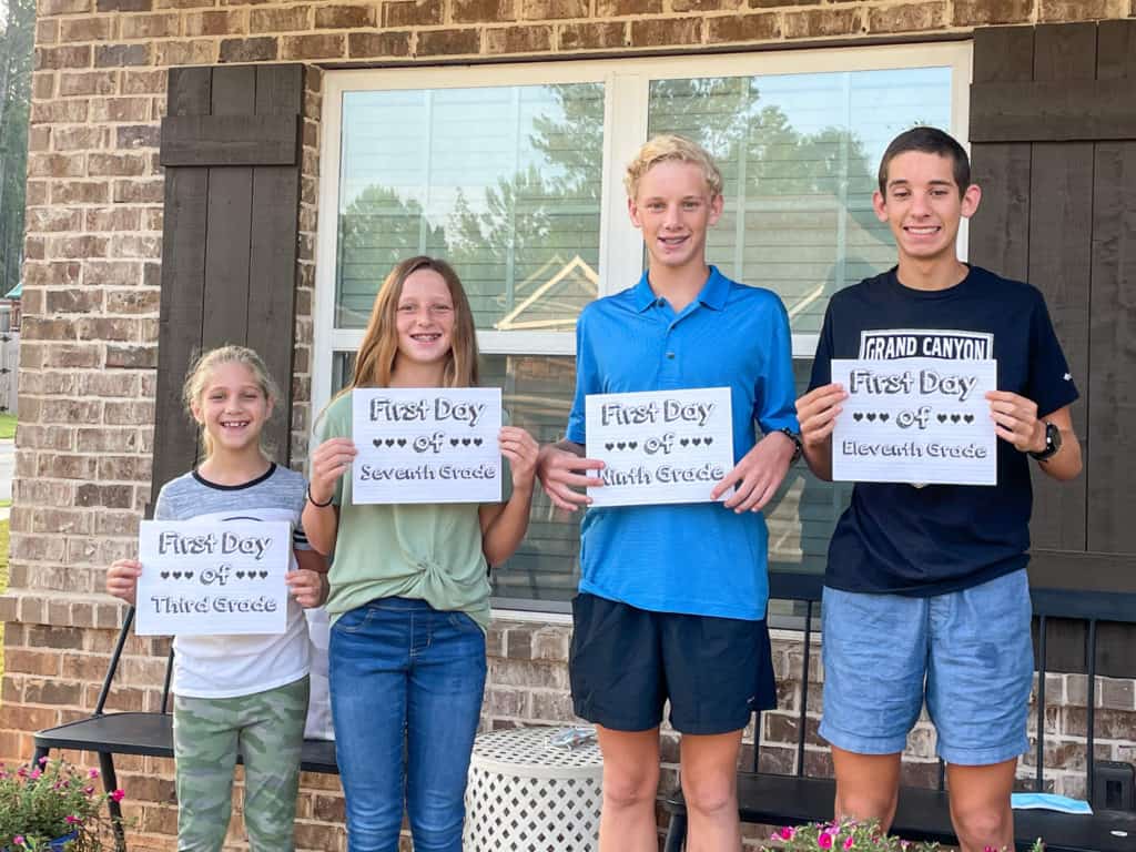 4 kids standing on front porch holding first day of school signs