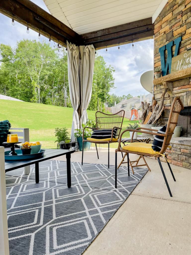 back porch curtains in the background of a back porch with grey and white rug, brown chairs with yellow seat cushions and a black outdoor coffee table. drop cloth curtains