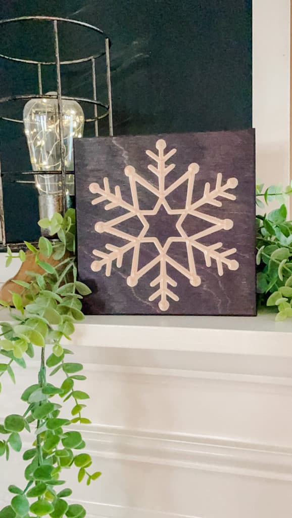 dark wood stained square with snowflake etched in the middle sitting on a mantel with greenery around it cottage keeping