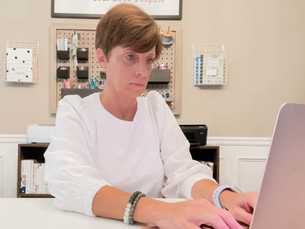 Short haired lady in white t-shirt with hands on the keyboard of a laptop with pegboard organization system in background.