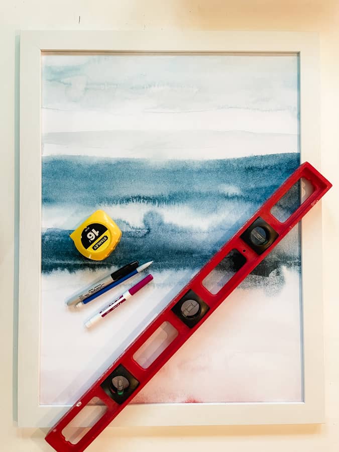 Supplies pictured for DIY Wall Calendar. white framed picture with blue and pink, red level, yellow tape measure and pens sitting on top of picture.