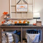 Fall themed Entry table with 3 black and white pillows on bottom left shelf and a basket with a black and white blanket on the bottom right shelf. Top shelf has a tiered tray with fall themed decor and orange flowers. Middle of table has silver flower holder containing orange flower blooms and white pumpkins. A lamp is on the right side of the table. Behind the table is a large white window with a fall themed framed print.