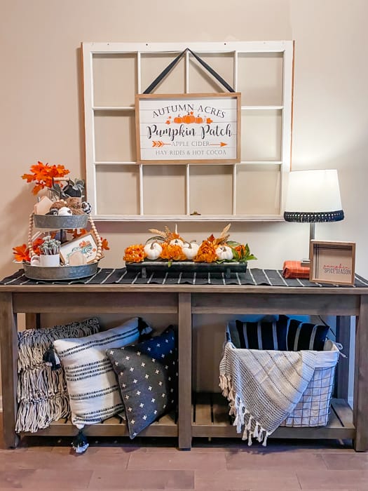 Fall themed Entry table with 3 black and white pillows on bottom left shelf and a basket with a black and white blanket on the bottom right shelf. Top shelf has a tiered tray with fall themed decor and orange flowers. Middle of table has silver flower holder containing orange flower blooms and white pumpkins. A lamp is on the right side of the table. Behind the table is a large white window with a fall themed framed print.
