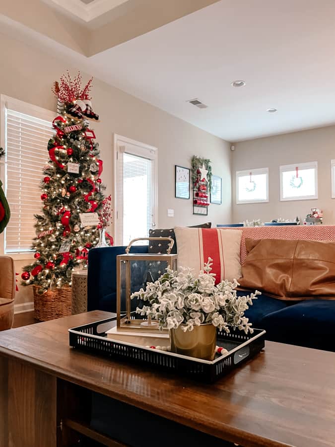 Cozy Christmas Living Room with wood table decorated with a black tray, gold accents and greenery. navy couch with leather pillow and red and white pillow. christmas tree decorated with red and white ribbon 