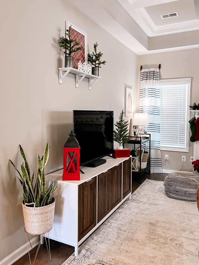 Cozy Christmas Living Room. snake plant in wicker planter. white and wood television stand with red lantern and christmas tree on red box on each side of a black flat screen tv. white wall shelf above tv decorated with christmas trees and framed red plaid paper