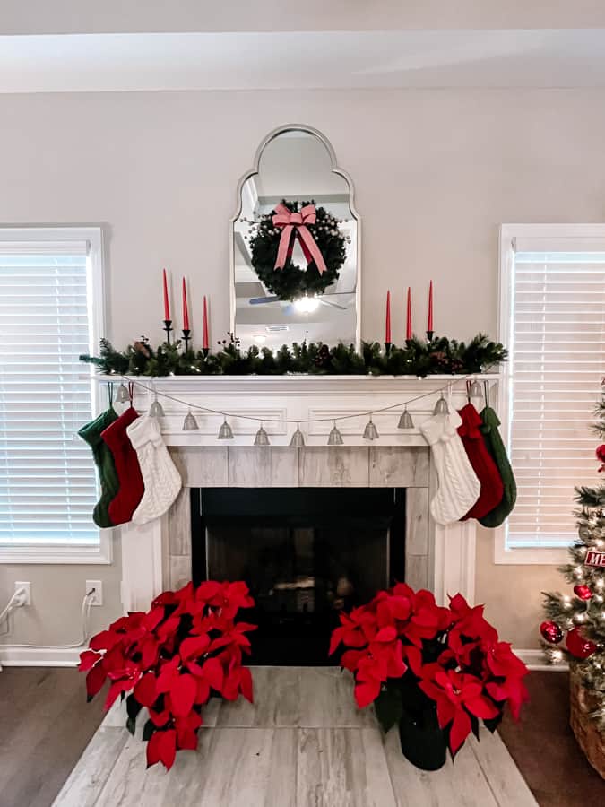 Cozy Christmas Living Room. white mantle decorated with greenery and red candles. a gold mirror in the center with a green wreath with red bow. red poinsettias on ground in front of fireplace. green red and white stockings and a bell garland