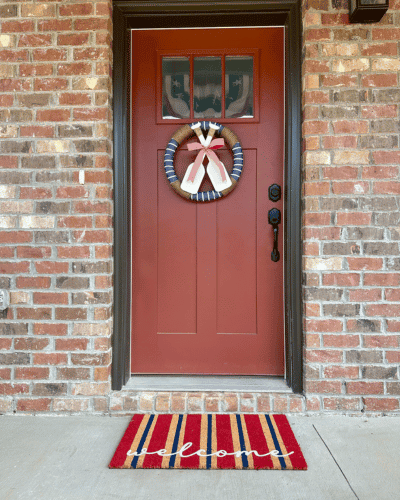 patriotic holiday decor on front porch. red and blue welcome mat in front of red door with blue and white round wreath with red bow on small white oars.