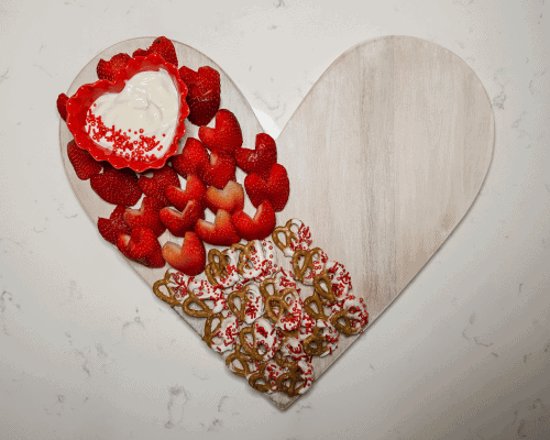 heart shaped wood board with heart shaped bowl containing white fruit dip. strawberry halves and white chocolate dipped pretzels on board.