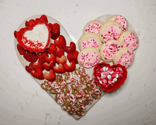valentine's charcuterie board on heart shaped, white board covered with strawberries, white chocolate pretzels and pink iced cookies