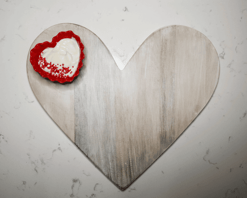 white wood heard shaped board with red heart shaped bowl containing white fruit dip