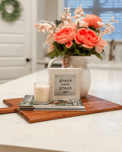 Valentine's Day Decorations on kitchen island with white countertop. Faux pink flowers in white ceramic pitcher. small white sign that says grace up on grace sitting beside a small white candle on a stack of magazines. 