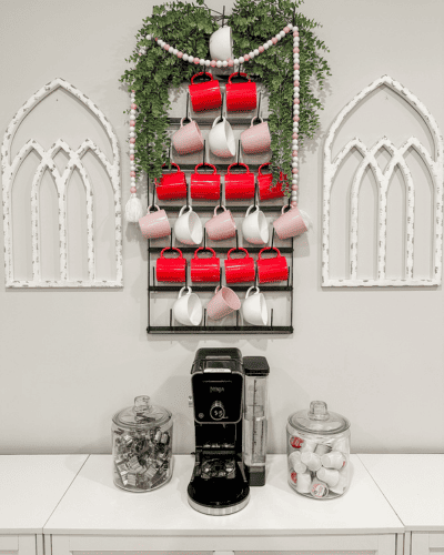 Valentine's Day Decor using a black metal cup rack with red, pink and white mugs hanging sporadically on the mug rack. Greenery and a pink and white bead garland draped over top of the mug rack.