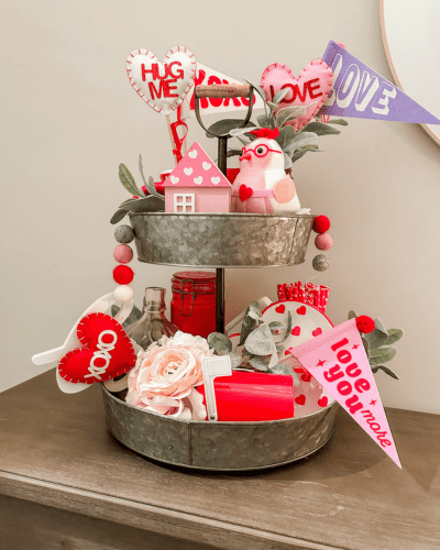 Valentine's Day decor in a galvanized metal tiered tray. tray has purple pennant that says LOVE, pink and white bird, pink wood house with white hearts on roof.