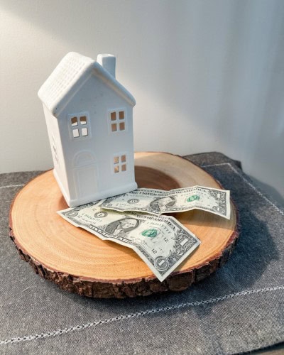 money saving tips picture with small white ceramic house sitting on a round wood charger with 2 $1 bills in front of it