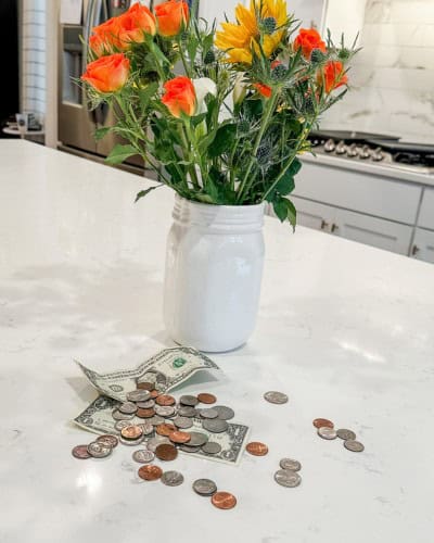 money saving tips picture with colorful flowers in a white vase. money is scattered on the counter top in front of the flower vase.