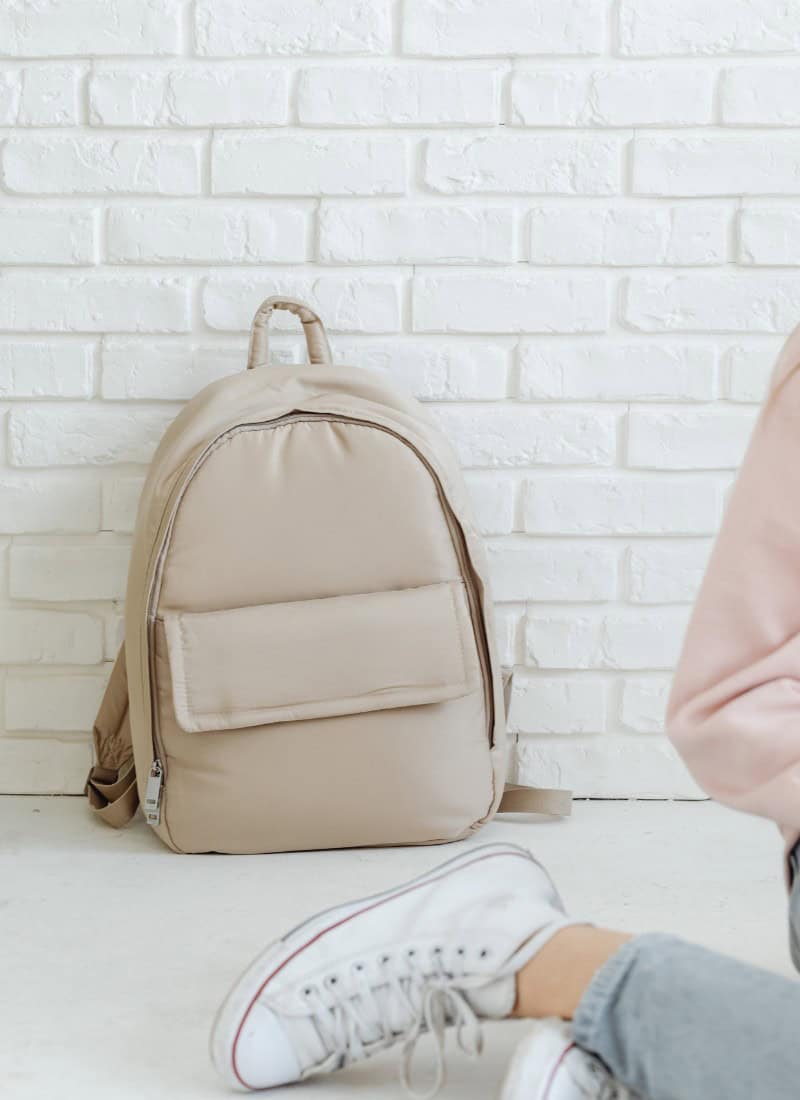 back to school cream backpack against a white brick wall.