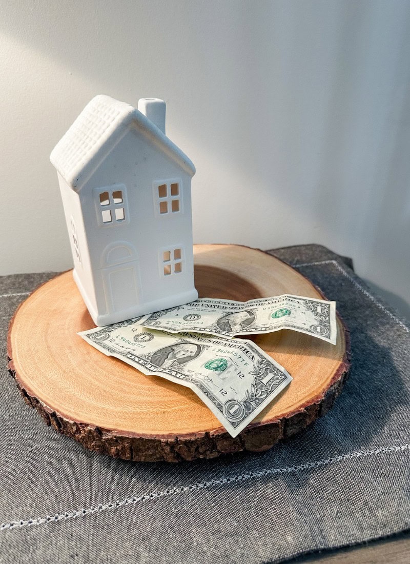 money saving tips. 2 $1 bills on a wood tree cutout with two small white houses sitting on top of it.