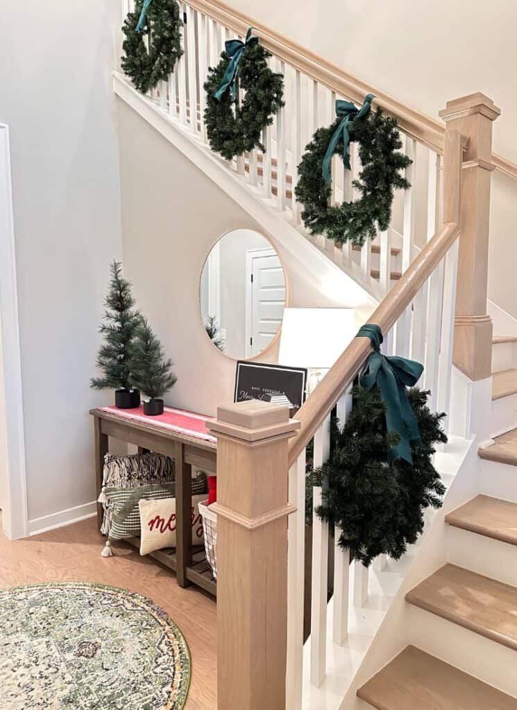 green wreaths hung on a white and light brown stair rail. Entry table with 2 green trees on a red table runner.