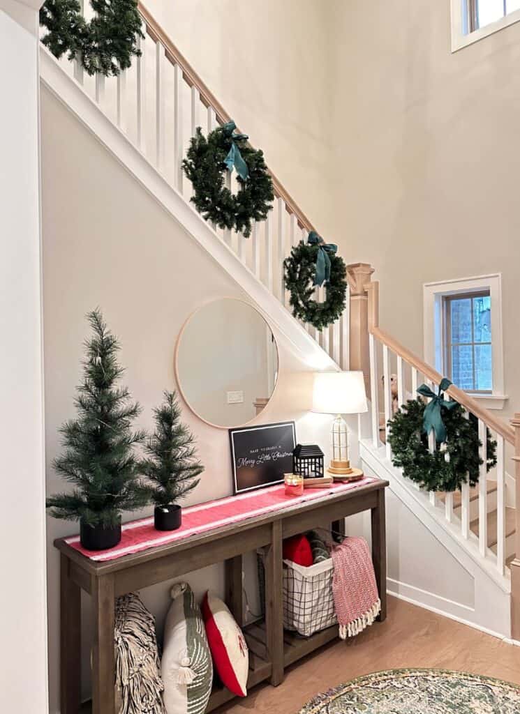 green wreaths hung on a white and light brown stair rail. Entry table with 2 green trees on a red table runner.