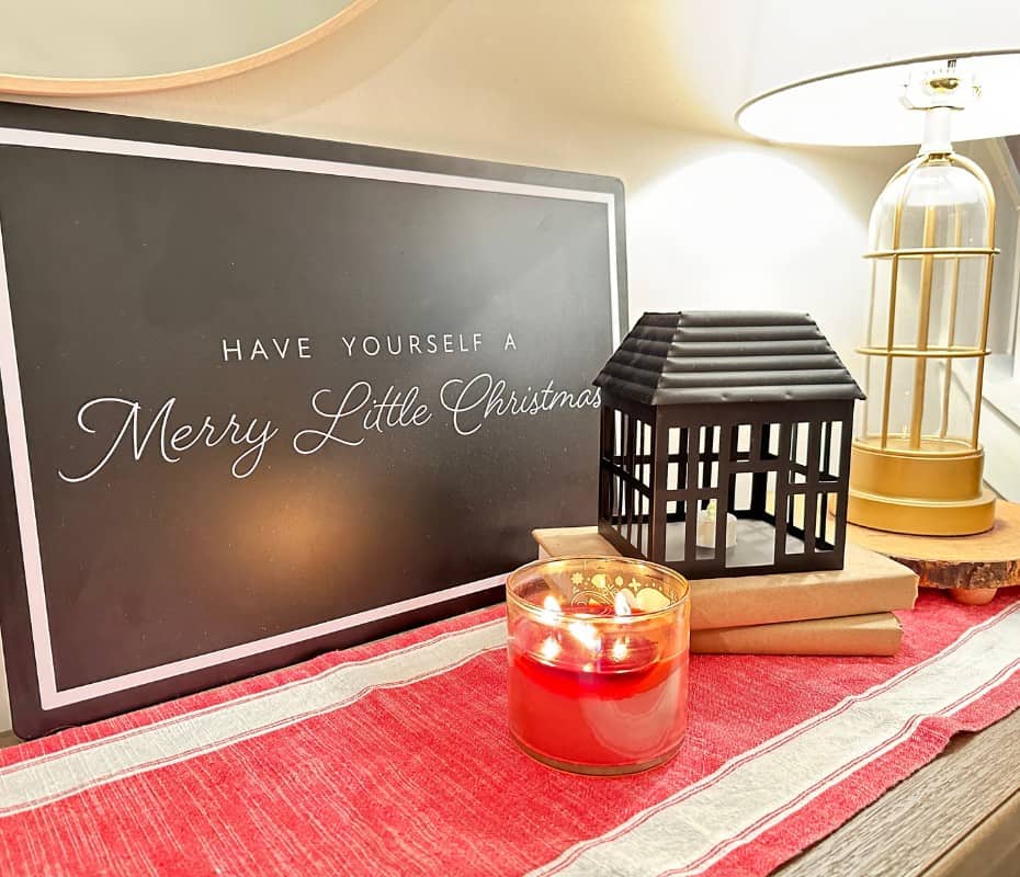 Black sign with white cursive Have Yourself a Merry Little Christmas on a red table runner with a lit candle and black metal house stacked on books.